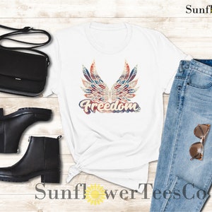 Puede incluir: Camiseta blanca con un gr&aacute;fico desgastado en rojo, blanco y azul de alas y la palabra "Freedom". La camiseta se combina con jeans azules, botas negras y un bolso bandolera negro.