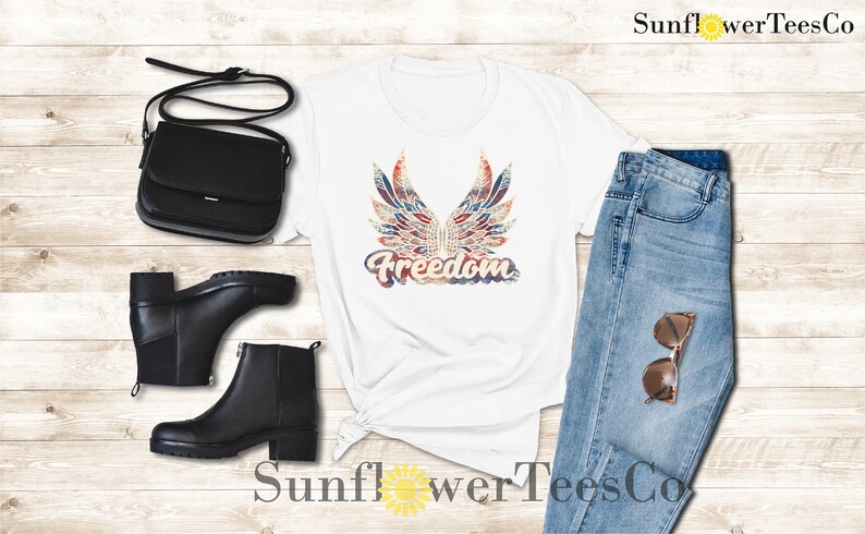 Puede incluir: Camiseta blanca con un gr&aacute;fico desgastado en rojo, blanco y azul de alas y la palabra "Freedom". La camiseta se combina con jeans azules, botas negras y un bolso bandolera negro.