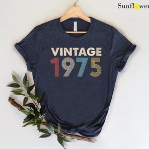May include: A navy blue t-shirt with the text "Vintage 1975" in a retro font with colorful stripes.