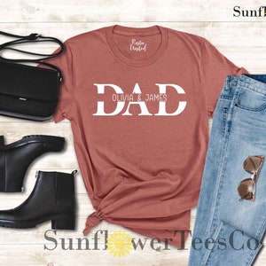 Op de afbeelding: Een roestkleurig T-shirt met het woord "DAD" in witte letters, met de namen "Olivia & James" eronder gedrukt. Het shirt wordt getoond op een houten oppervlak met een zwarte tas, zwarte laarzen en een blauwe jeans.