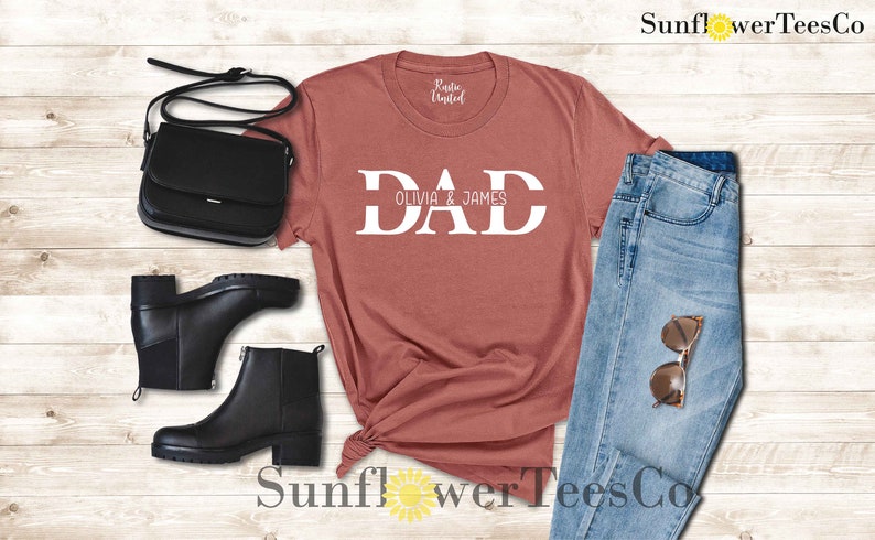 Op de afbeelding: Een roestkleurig T-shirt met het woord "DAD" in witte letters, met de namen "Olivia & James" eronder gedrukt. Het shirt wordt getoond op een houten oppervlak met een zwarte tas, zwarte laarzen en een blauwe jeans.