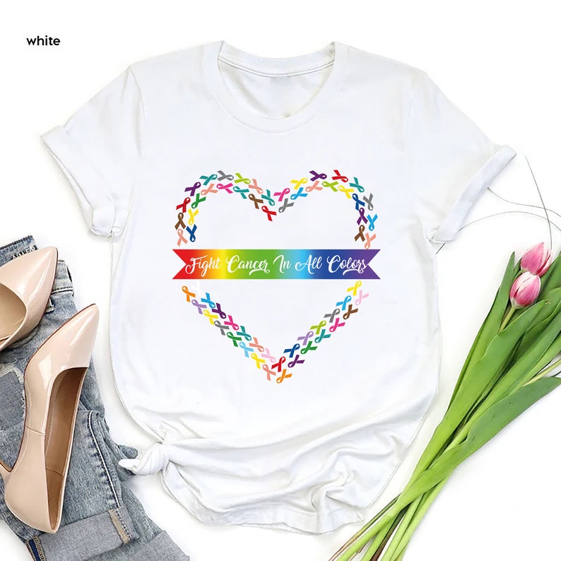 May include: White t-shirt featuring a heart-shaped design made of colorful ribbons. The shirt has the text "Fight Cancer In All Colors" across the center. The shirt is a crew neck and has short sleeves.