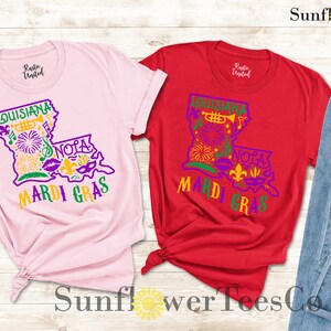 May include: Two t-shirts with a graphic design of the state of Louisiana with Mardi Gras themed icons. The shirts are pink and red. The text on the shirts says "Louisiana" and "Mardi Gras".