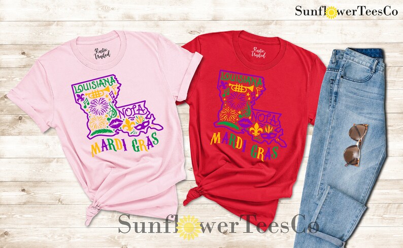 May include: Two t-shirts with a graphic design of the state of Louisiana with Mardi Gras themed icons. The shirts are pink and red. The text on the shirts says "Louisiana" and "Mardi Gras".