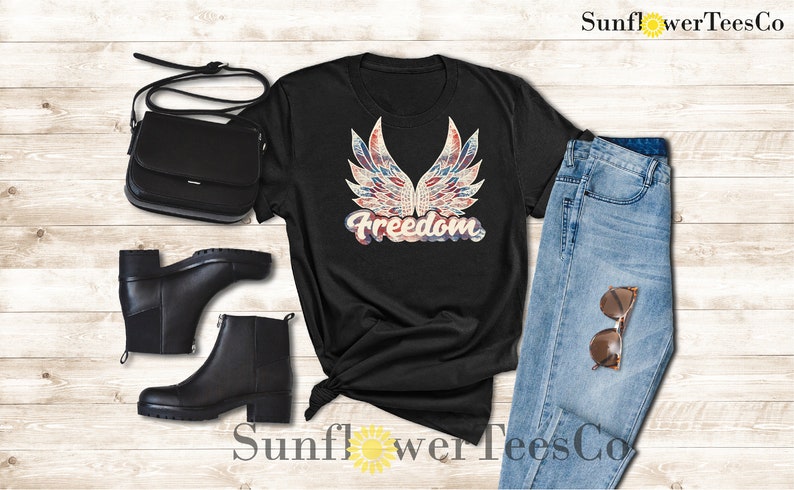 Puede incluir: Camiseta negra con un dise&ntilde;o estampado de alas en rojo, blanco y azul y la palabra "Freedom". La camiseta se muestra con un par de botas negras, un bolso negro y unos vaqueros azules.