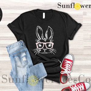 May include: Black t-shirt with a white graphic of a bunny wearing pink glasses. The t-shirt is laid flat on a wooden surface with a pair of blue jeans and red sneakers.