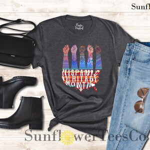 May include: A dark gray t-shirt with a graphic of raised fists and the text "Hispanic Heritage Month". The graphic is colorful and features a stylized map of the United States.