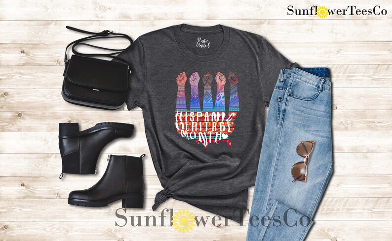 May include: A dark gray t-shirt with a graphic of raised fists and the text "Hispanic Heritage Month". The graphic is colorful and features a stylized map of the United States.