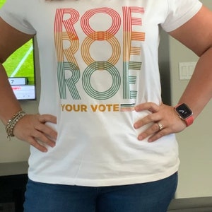 K&ouml;nnte beinhalten: Wei&szlig;es T-Shirt mit dem Text "ROE YOUR VOTE" in einem vertikalen Retro-Design. Das Wort "ROE" ist dreimal gestapelt, jeweils in einer anderen Farbe: Rot, Orange und Gr&uuml;n. Das Shirt wird mit einer blauen Jeans kombiniert.