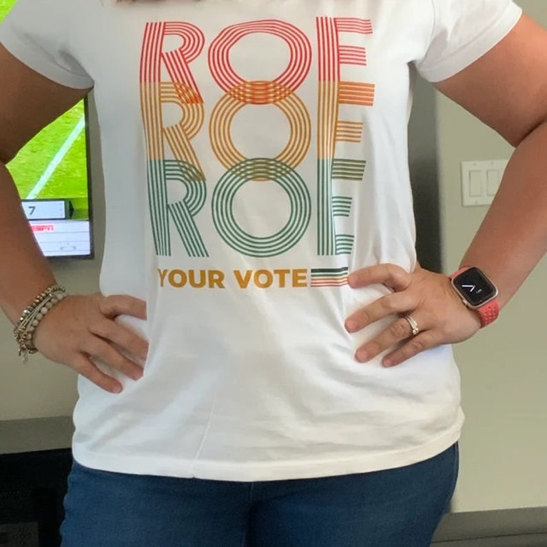 K&ouml;nnte beinhalten: Wei&szlig;es T-Shirt mit dem Text "ROE YOUR VOTE" in einem vertikalen Retro-Design. Das Wort "ROE" ist dreimal gestapelt, jeweils in einer anderen Farbe: Rot, Orange und Gr&uuml;n. Das Shirt wird mit einer blauen Jeans kombiniert.