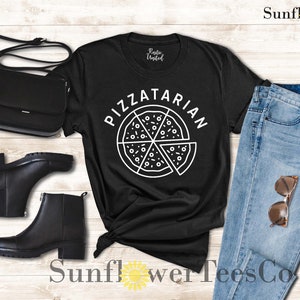May include: Black t-shirt with a white graphic of a pizza slice and the text "PIZZATARIAN". The shirt is laid out on a wooden surface with a pair of black boots, a black purse, and blue jeans.