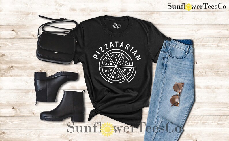 May include: Black t-shirt with a white graphic of a pizza slice and the text "PIZZATARIAN". The shirt is laid out on a wooden surface with a pair of black boots, a black purse, and blue jeans.