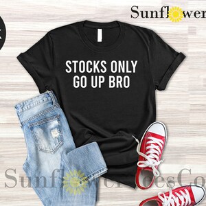May include: Black t-shirt with the text "Stocks Only Go Up Bro" in white lettering. The shirt is laid flat on a wooden surface with a pair of blue jeans and red sneakers.