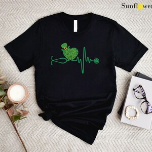 May include: Black t-shirt with a green shamrock heart and a stethoscope, representing a heartbeat. The shamrock heart has a green leprechaun hat on top. The text "SunflwerTeesCo" is in the top right corner of the image.