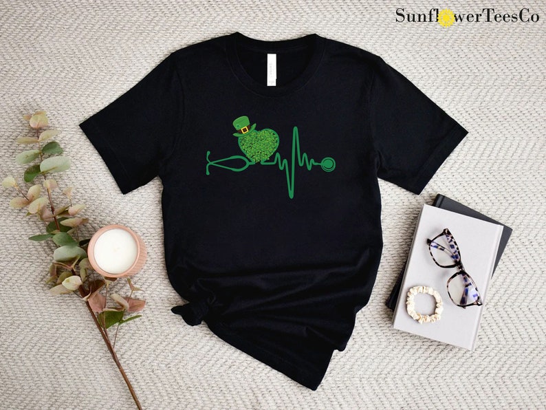 May include: Black t-shirt with a green shamrock heart and a stethoscope, representing a heartbeat. The shamrock heart has a green leprechaun hat on top. The text "SunflwerTeesCo" is in the top right corner of the image.