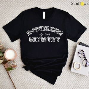 May include: Black t-shirt with white text that reads "Motherhood is my Ministry" in a varsity style font.