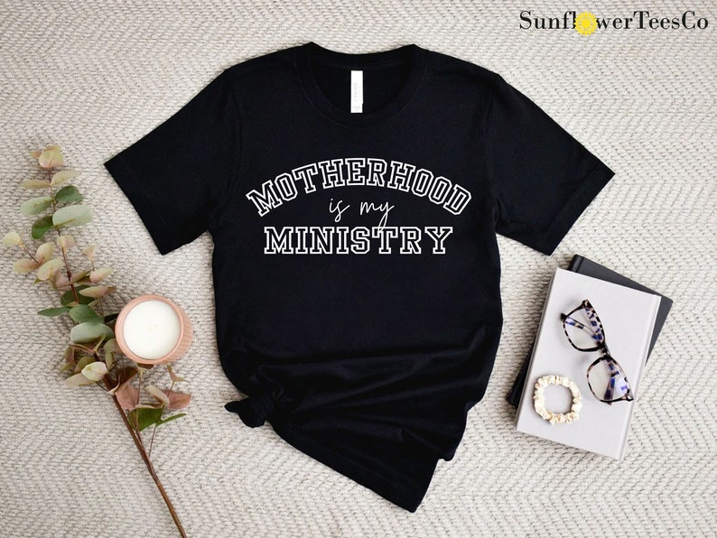 May include: Black t-shirt with white text that reads "Motherhood is my Ministry" in a varsity style font.
