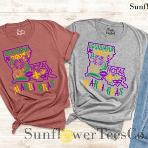 May include: Two t-shirts with a graphic design of the state of Louisiana with the words "Louisiana Mardi Gras" and "NOLA" in purple and gold. The shirts are on a wooden background with a pair of blue jeans and sunglasses.