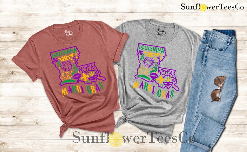 May include: Two t-shirts with a graphic design of the state of Louisiana with the words "Louisiana Mardi Gras" and "NOLA" in purple and gold. The shirts are on a wooden background with a pair of blue jeans and sunglasses.