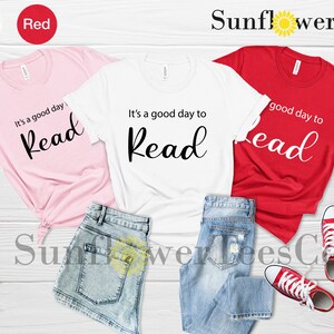 Op de afbeelding: Drie t-shirts in roze, wit en rood, elk met de tekst "It's a good day to Read" in zwart script. De shirts worden getoond met blauwe spijkerbroekshorts en rode canvas sneakers op een wit houten oppervlak.
