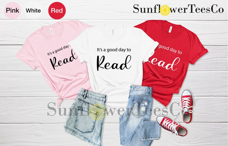 Op de afbeelding: Drie t-shirts in roze, wit en rood, elk met de tekst "It's a good day to Read" in zwart script. De shirts worden getoond met blauwe spijkerbroekshorts en rode canvas sneakers op een wit houten oppervlak.