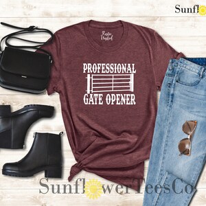 Op de afbeelding: Een maroon t-shirt met de tekst "Professional Gate Opener" en een wit silhouet van een poort. Het shirt wordt gecombineerd met een blauwe jeans, zwarte laarzen en een zwarte handtas.