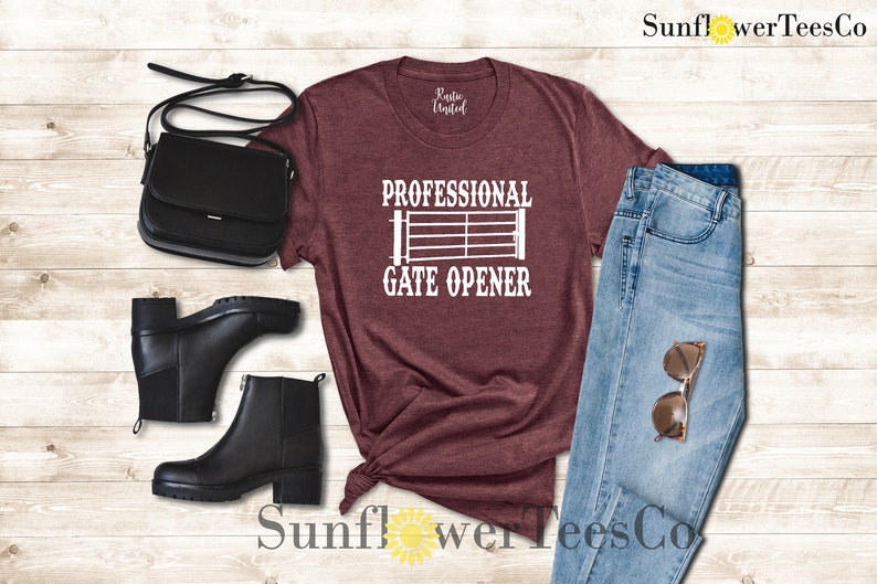 Op de afbeelding: Een maroon t-shirt met de tekst "Professional Gate Opener" en een wit silhouet van een poort. Het shirt wordt gecombineerd met een blauwe jeans, zwarte laarzen en een zwarte handtas.