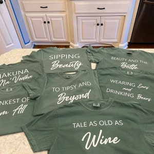 K&ouml;nnte beinhalten: Mehrere olivgr&uuml;ne T-Shirts mit wei&szlig;em Text. Die Shirts tragen Aufschriften wie "Sipping Beauty", "To Tipsy and Beyond" und "Tale as Old as Wine". Die Shirts liegen auf einer hellen Arbeitsplatte.