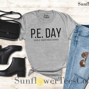 Op de afbeelding: Grijs T-shirt met de tekst "P.E. DAY (aka best day ever)" erop gedrukt. Het shirt wordt gecombineerd met een blauwe jeans, zwarte laarzen en een zwarte handtas.