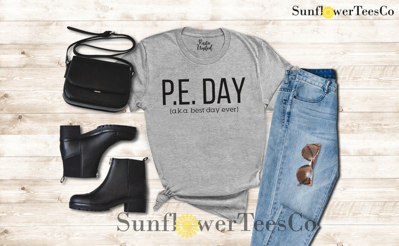 Op de afbeelding: Grijs T-shirt met de tekst "P.E. DAY (aka best day ever)" erop gedrukt. Het shirt wordt gecombineerd met een blauwe jeans, zwarte laarzen en een zwarte handtas.