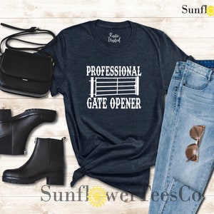 Op de afbeelding: Een donkerblauw t-shirt met de tekst "Professional Gate Opener" en een wit silhouet van een poort. Het t-shirt wordt getoond met een paar zwarte enkellaarsjes, een zwarte handtas en een blauwe jeans.