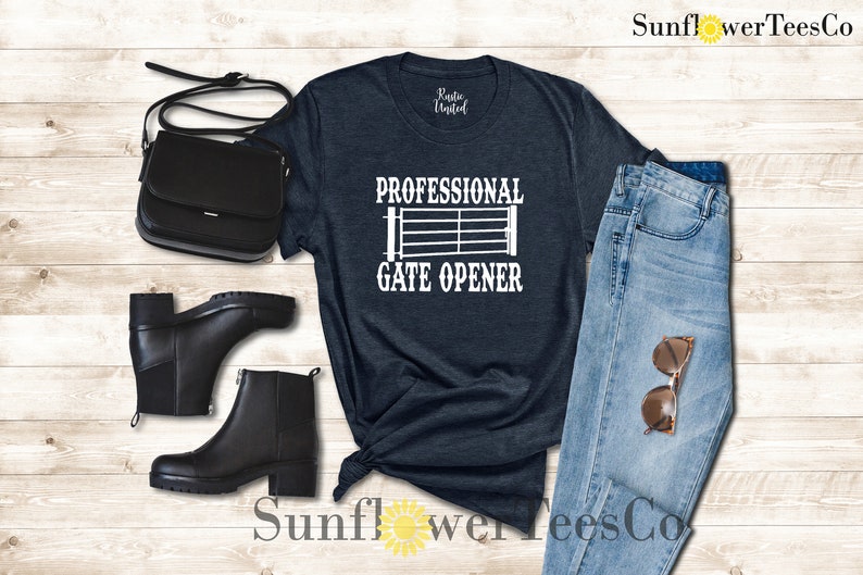 Op de afbeelding: Een donkerblauw t-shirt met de tekst "Professional Gate Opener" en een wit silhouet van een poort. Het t-shirt wordt getoond met een paar zwarte enkellaarsjes, een zwarte handtas en een blauwe jeans.