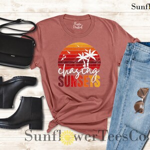 May include: A maroon t-shirt with a vintage sunset graphic and the text "chasing sunsets". The shirt is paired with blue jeans, black ankle boots, and a black crossbody bag.