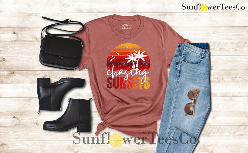 May include: A maroon t-shirt with a vintage sunset graphic and the text "chasing sunsets". The shirt is paired with blue jeans, black ankle boots, and a black crossbody bag.