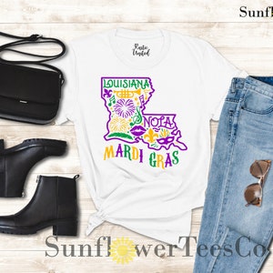 May include: White t-shirt with a purple and yellow graphic of the state of Louisiana with the words "Louisiana" and "Mardi Gras". The graphic includes symbols like a trumpet, fleur-de-lis, and a mask. The shirt is paired with blue jeans, a black purse, and black ankle boots.