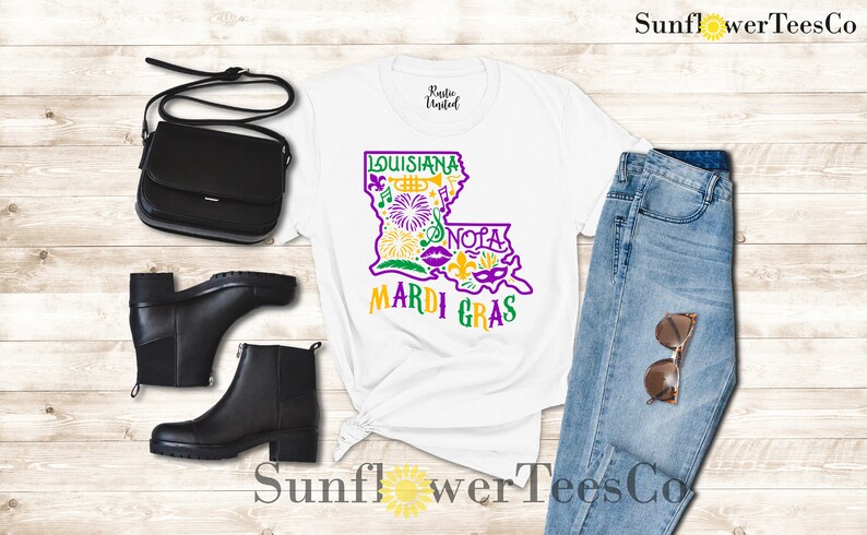 May include: White t-shirt with a purple and yellow graphic of the state of Louisiana with the words "Louisiana" and "Mardi Gras". The graphic includes symbols like a trumpet, fleur-de-lis, and a mask. The shirt is paired with blue jeans, a black purse, and black ankle boots.