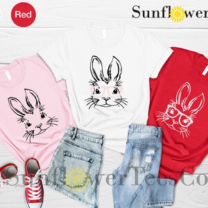 May include: Three t-shirts with a graphic of a bunny wearing glasses. The shirts are pink, white, and red. The text "Sunflower TeesCo" is visible at the top of the image.