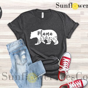 May include: A dark heather grey t-shirt with a white silhouette of a mama bear with floral details and the word "Mama" inside the bear. The shirt is laid flat on a wooden surface with a pair of red sneakers and blue jeans.