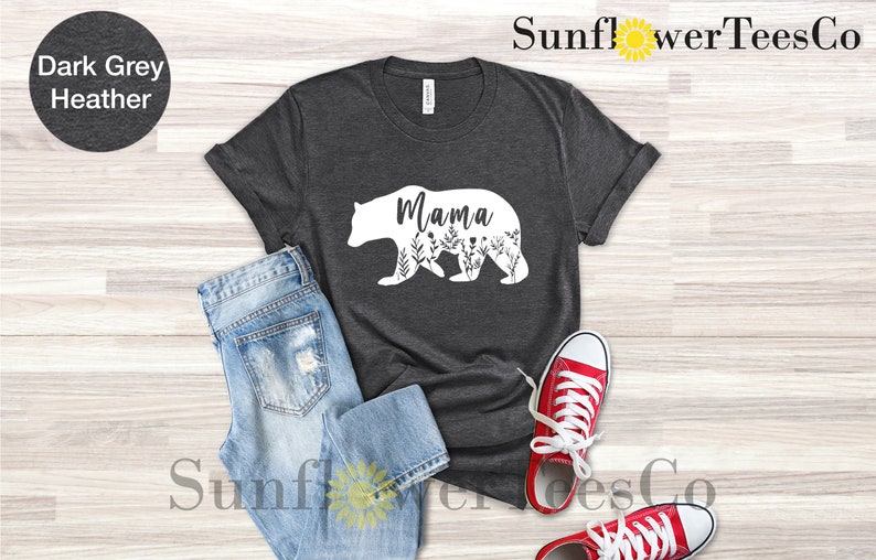 May include: A dark heather grey t-shirt with a white silhouette of a mama bear with floral details and the word "Mama" inside the bear. The shirt is laid flat on a wooden surface with a pair of red sneakers and blue jeans.