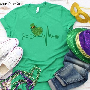 May include: Green t-shirt with a green shamrock heart and a stethoscope, a heartbeat line, and a stethoscope. The shirt is on a white wooden background with green, gold, and purple Mardi Gras beads, a gold mask, and a green, gold, and purple sequined hat.