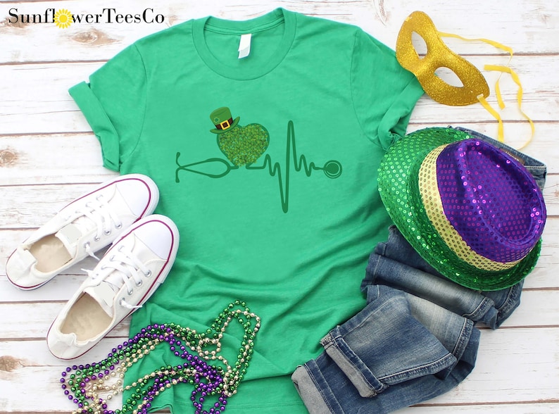 May include: Green t-shirt with a green shamrock heart and a stethoscope, a heartbeat line, and a stethoscope. The shirt is on a white wooden background with green, gold, and purple Mardi Gras beads, a gold mask, and a green, gold, and purple sequined hat.