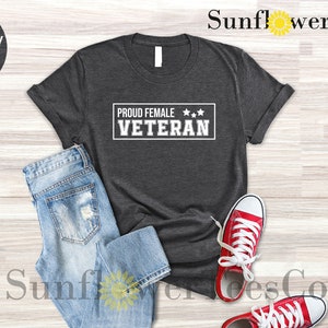May include: Dark grey heather t-shirt with the text "Proud Female Veteran" in white with three stars above the text. The shirt is laid flat on a light wood surface with a pair of blue jeans and red sneakers.