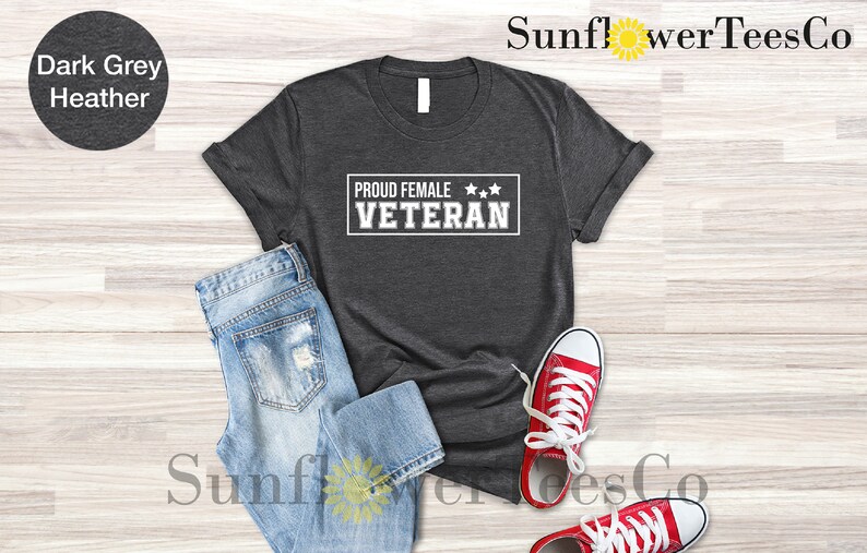May include: Dark grey heather t-shirt with the text "Proud Female Veteran" in white with three stars above the text. The shirt is laid flat on a light wood surface with a pair of blue jeans and red sneakers.
