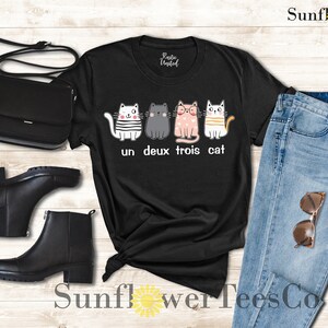 以下が含まれることがあります： 4匹の猫が並んだグラフィックがプリントされた黒いTシャツ。猫はそれぞれ違う服を着ていて、表情も違います。「un deux trois cat」という文字が猫の下にプリントされています。