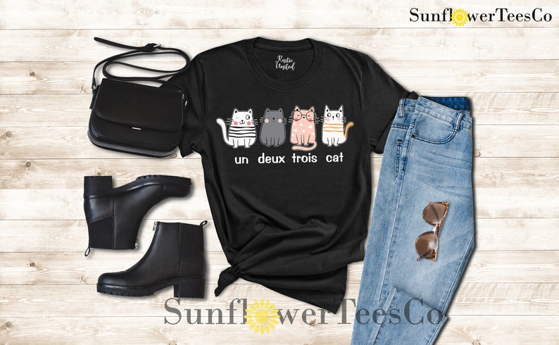 以下が含まれることがあります： 4匹の猫が並んだグラフィックがプリントされた黒いTシャツ。猫はそれぞれ違う服を着ていて、表情も違います。「un deux trois cat」という文字が猫の下にプリントされています。