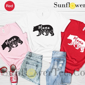 May include: Three t-shirts with a bear silhouette design. The bear is outlined in black on a pink shirt, white on a white shirt, and black on a red shirt. The word "Mama" is inside the bear silhouette. The shirts are laid out on a wooden surface with denim shorts.