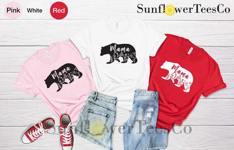 May include: Three t-shirts with a bear silhouette design. The bear is outlined in black on a pink shirt, white on a white shirt, and black on a red shirt. The word "Mama" is inside the bear silhouette. The shirts are laid out on a wooden surface with denim shorts.