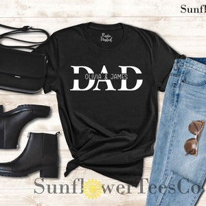 Op de afbeelding: Zwart T-shirt met witte tekst die "DAD Olivia & James" zegt. Het shirt ligt op een houten oppervlak met een paar zwarte laarzen, een zwarte tas en een blauwe jeans.