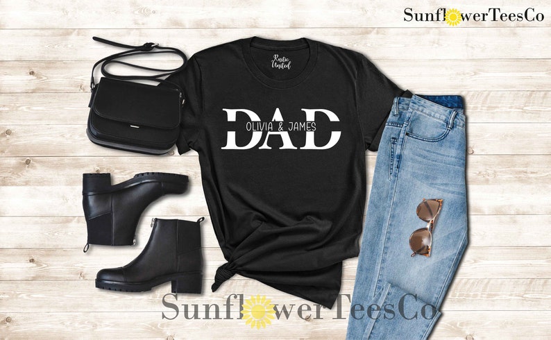 Op de afbeelding: Zwart T-shirt met witte tekst die "DAD Olivia & James" zegt. Het shirt ligt op een houten oppervlak met een paar zwarte laarzen, een zwarte tas en een blauwe jeans.