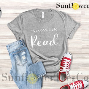 Op de afbeelding: Lichtgrijs heather t-shirt met de tekst "It's a good day to Read" in witte letters. Het shirt wordt gecombineerd met lichtblauwe jeans en rode canvas sneakers met witte veters. De afbeelding is een flat lay.